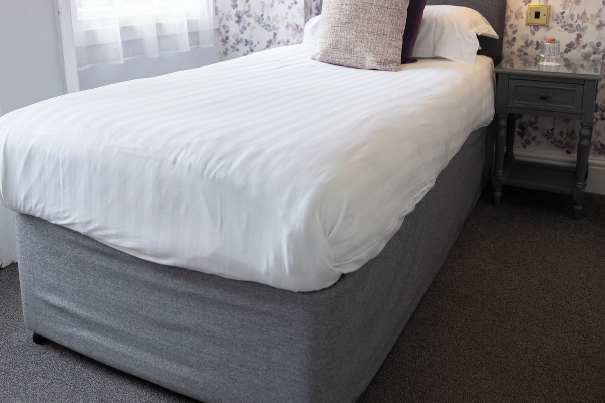 Un dormitorio con cama, mesita de noche, lámpara, ventana con cortinas y papel tapiz con motivos florales.