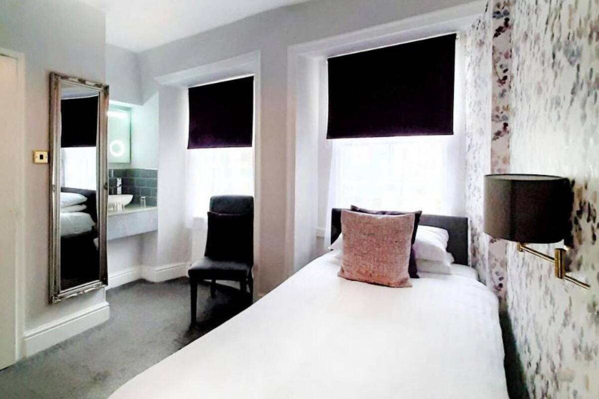 Habitación de hotel con cama, una silla, un espejo, una ventana con cortina opaca y una lámpara de pared.