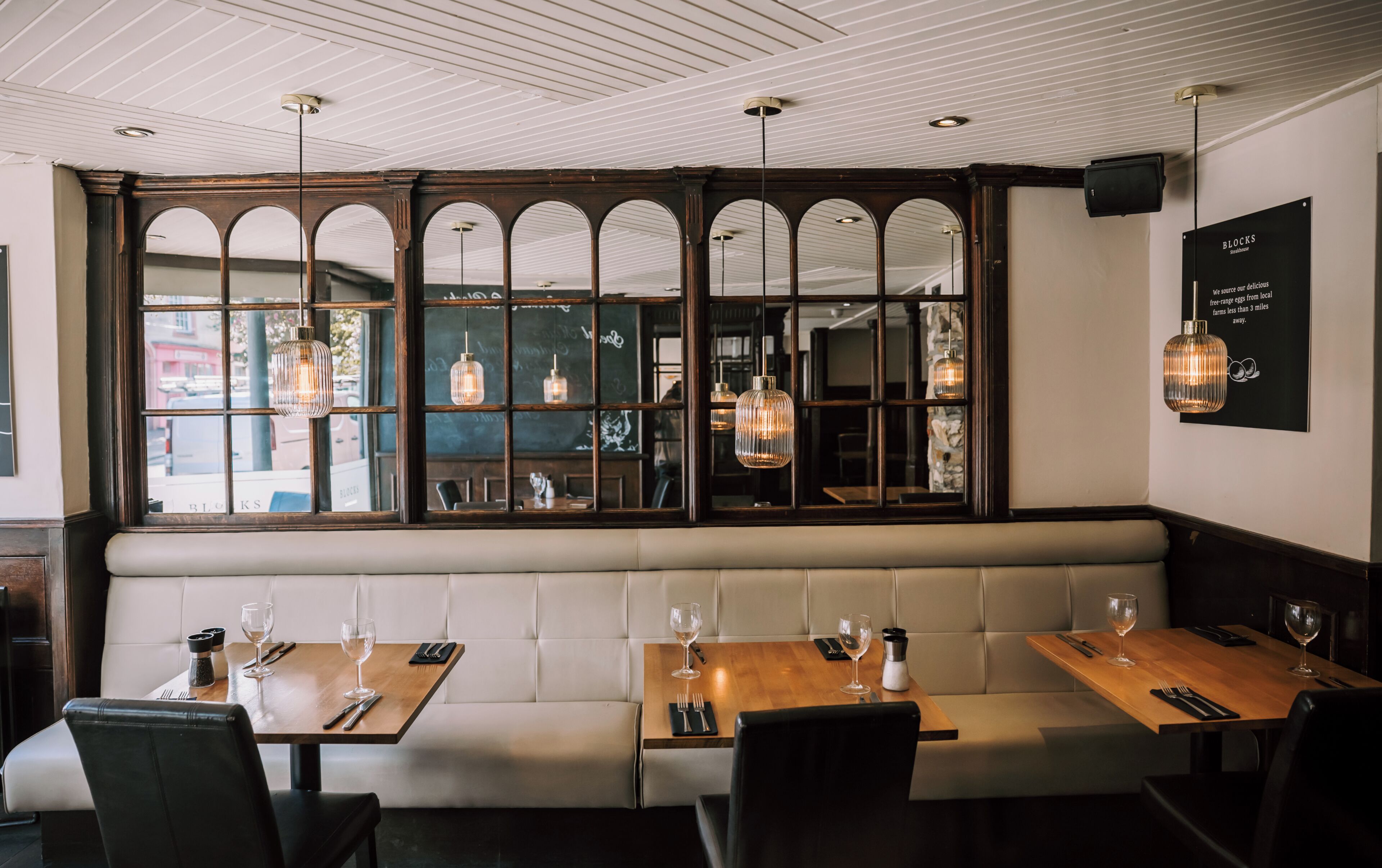 Interior de restaurante con techo alto, paneles de madera y un amplio ventanal con múltiples divisiones. Las mesas están dispuestas con copas de vino y cubiertos.