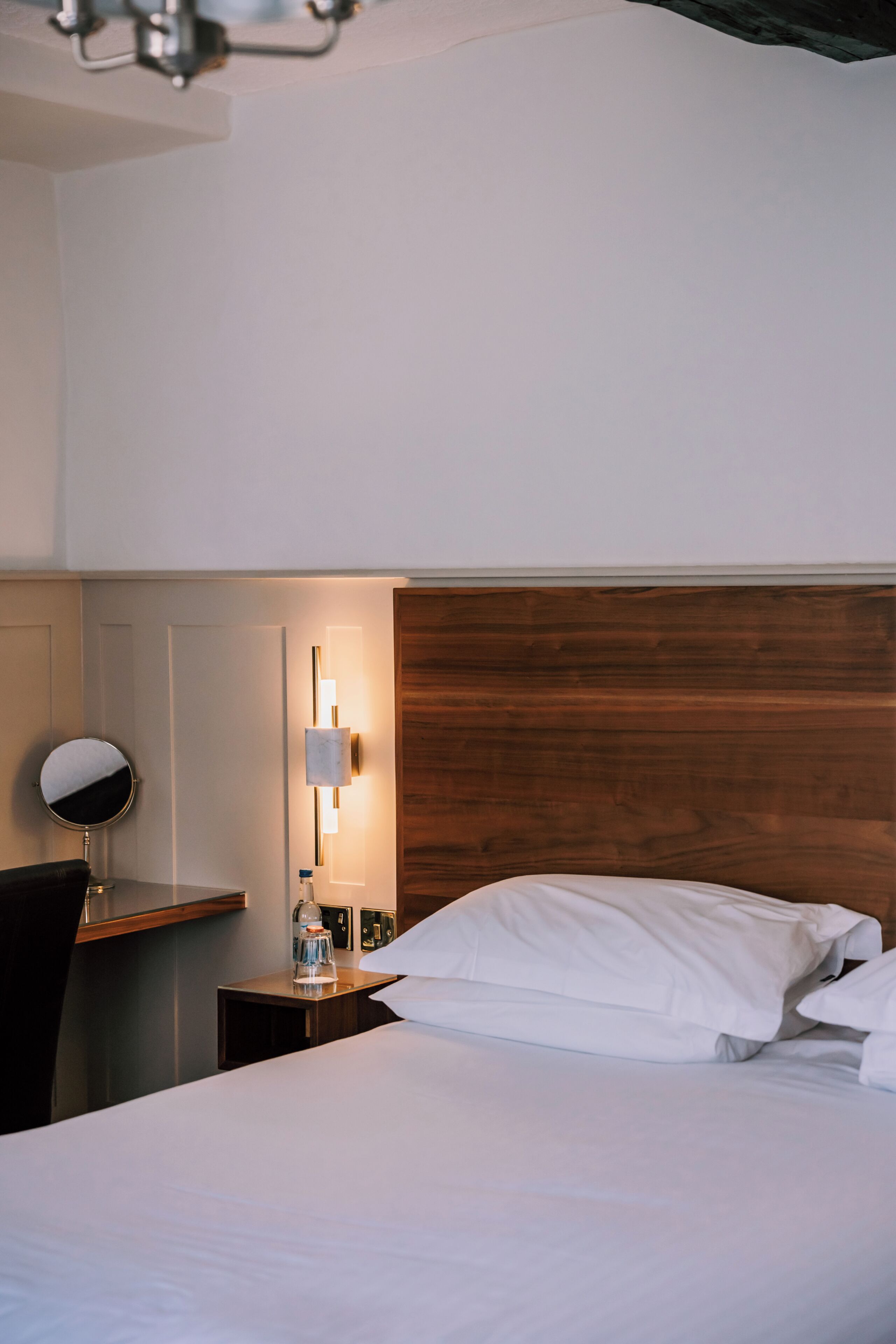 A hotel room with a wooden headboard, a round mirror, a bedside table with a lamp, and a neatly made bed.
