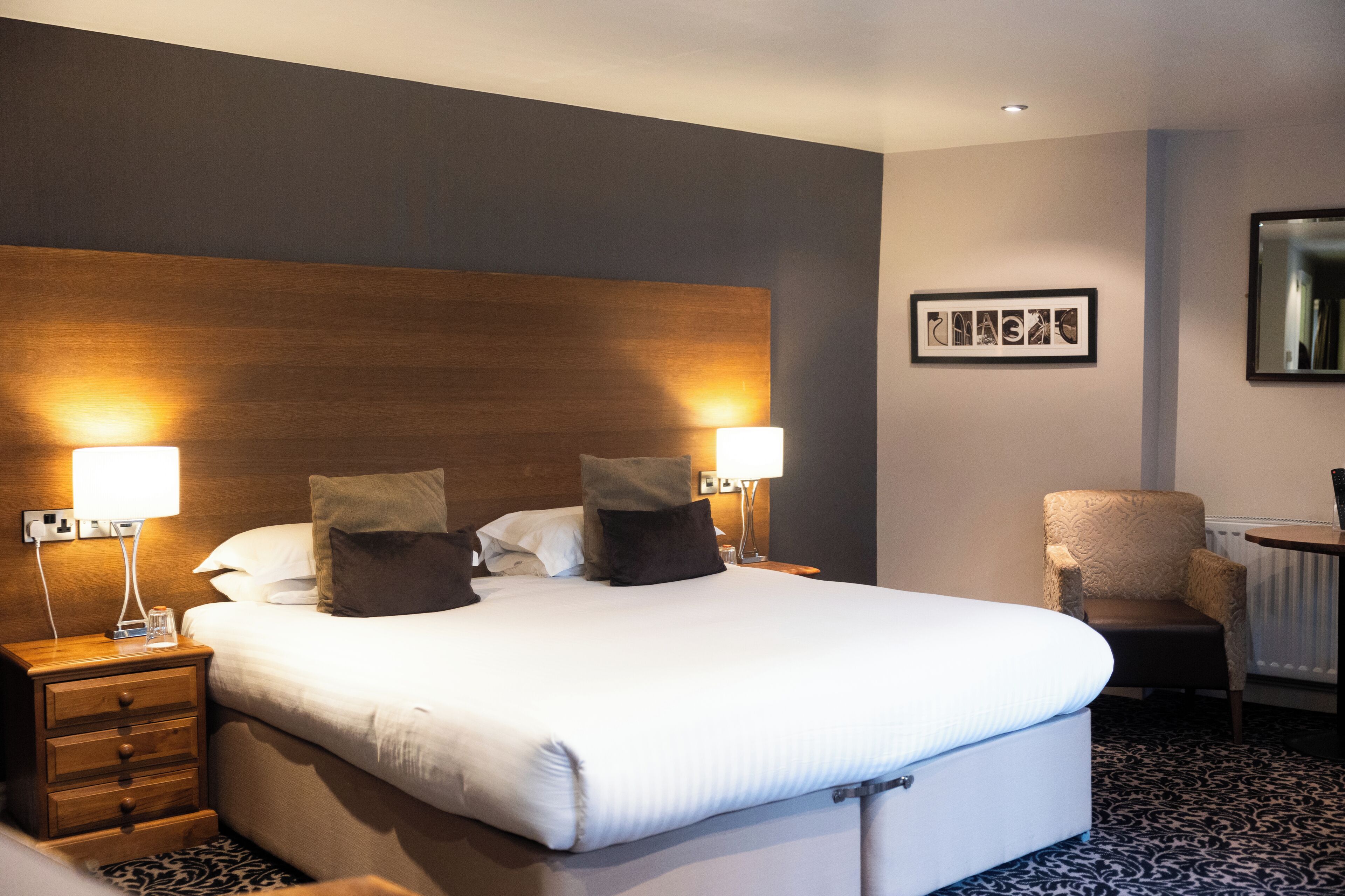 A hotel room with a large bed, two bedside lamps, a chair, and a framed picture on the wall.