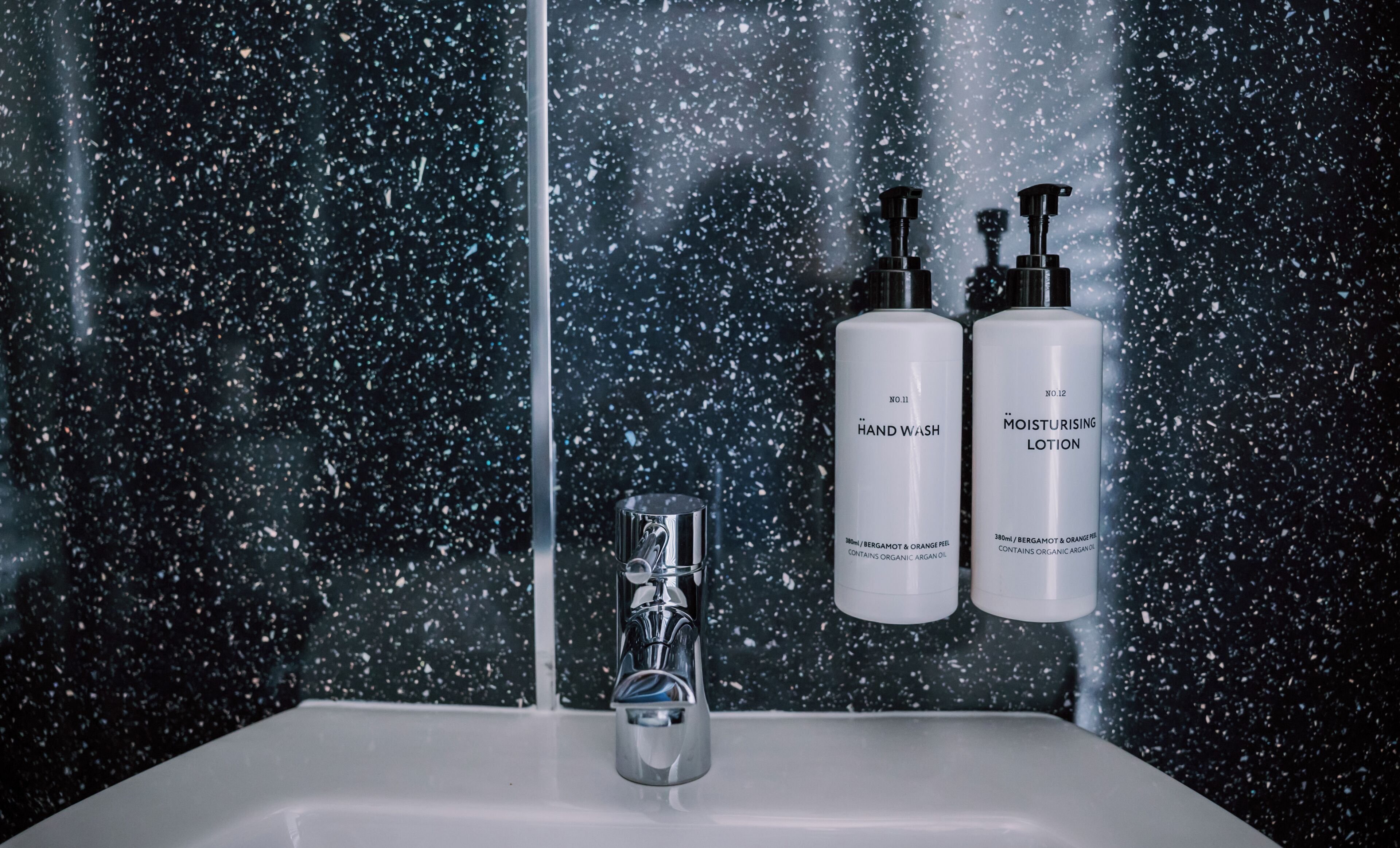 A bathroom with a sink, two bottles of hand wash and moisturizing lotion, and a faucet.