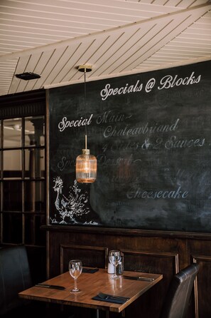 A restaurant interior with a chalkboard menu listing specials, a wooden table with a wine glass, and a hanging light fixture. - The Allerdale Court Hotel (Cockermouth)