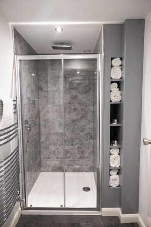 A walk-in shower with a glass door, a towel rack, and a shelf with towels. - The Allerdale Court Hotel (Cockermouth)