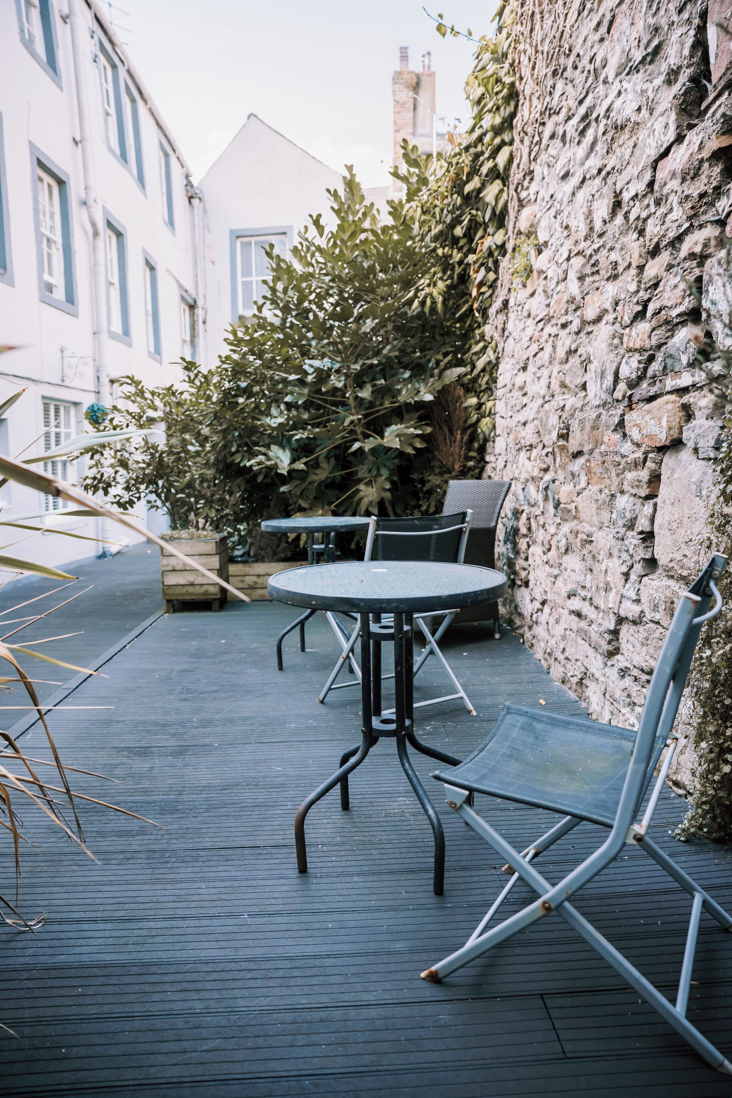 An outdoor seating area with a round table and chairs, a stone wall, and a building in the background.