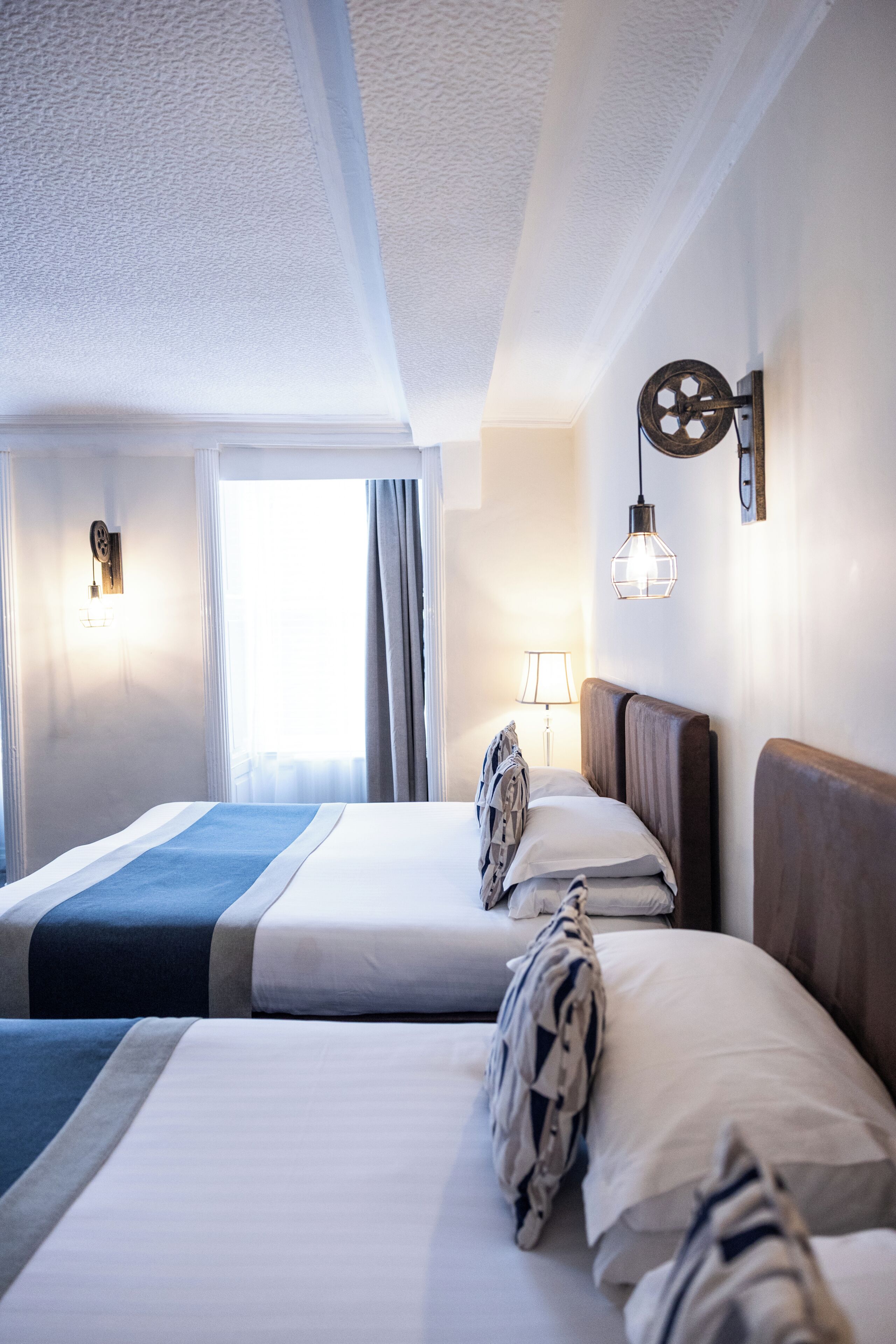 A hotel room with two beds, a window with curtains, a wall-mounted lamp, and a ceiling with a textured finish.