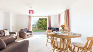 Ein modernes Wohnzimmer mit einer Couch, einem Esstisch mit Stühlen, einem Fernseher und Blick auf einen Garten durch große Fenster.