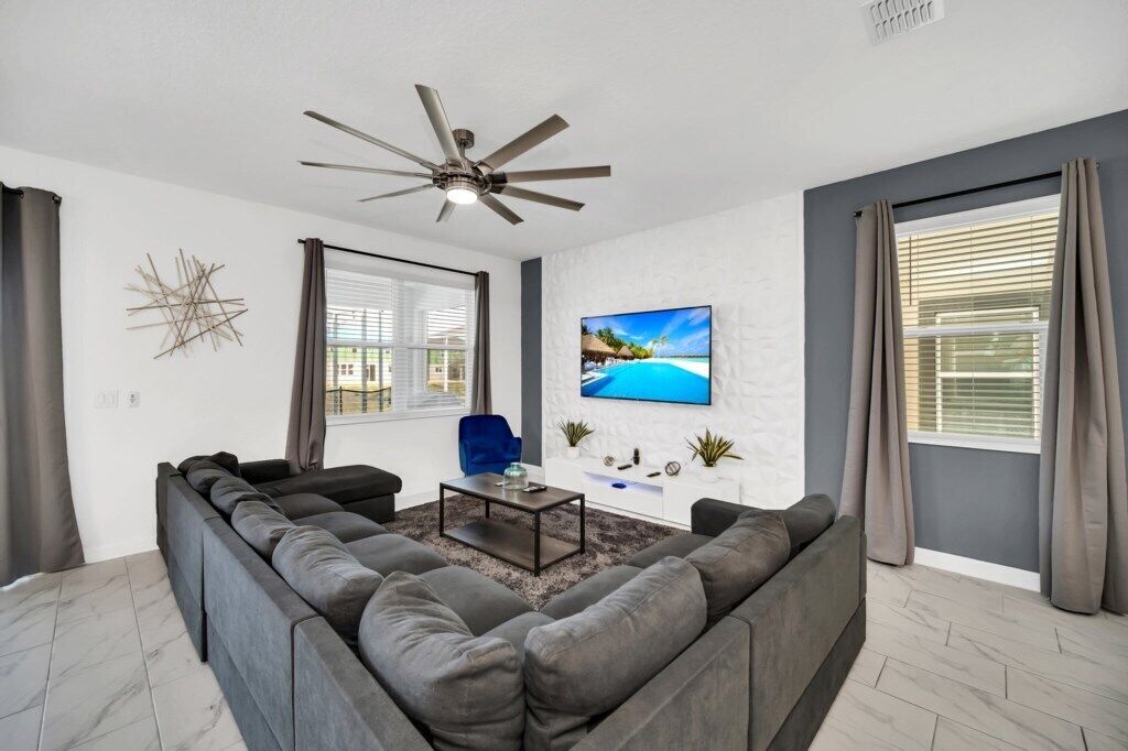 A modern living room with a sectional sofa, a coffee table, a ceiling fan, and a TV mounted on the wall.