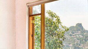 Una habitación con un ventanal grande, dos sillones grises, una mesita con una vela y un florero con flores, y vistas a un lago y colinas.
