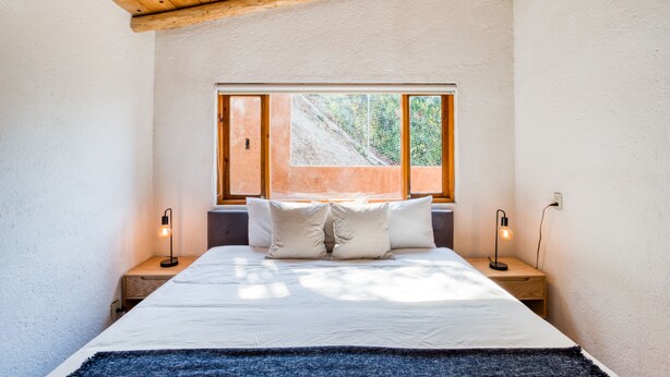Un dormitorio con techo de madera, un ventanal grande, una cama con ropa de cama blanca y dos lámparas de noche.