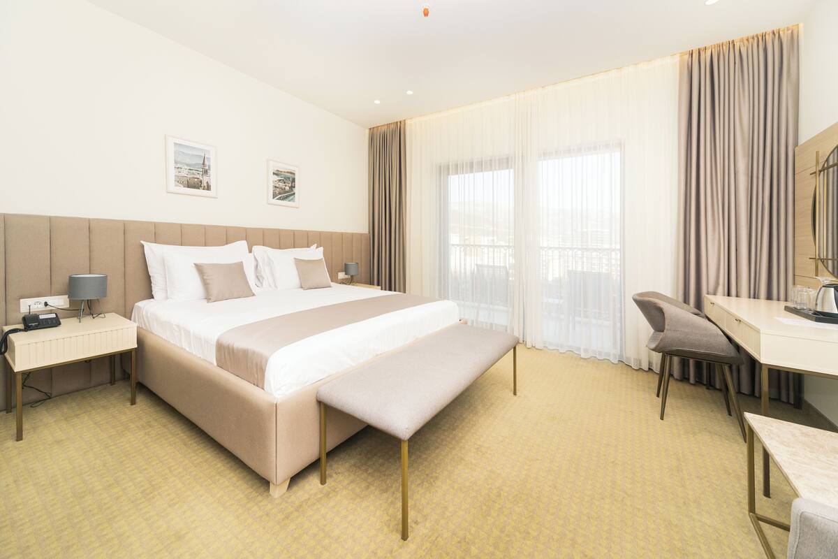 A hotel room with a large bed, a desk, a chair, a bench, and a window with curtains.