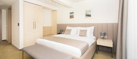 Quarto de hotel com uma cama grande, mesa de cabeceira com abajur, um banco e guarda-roupa.
