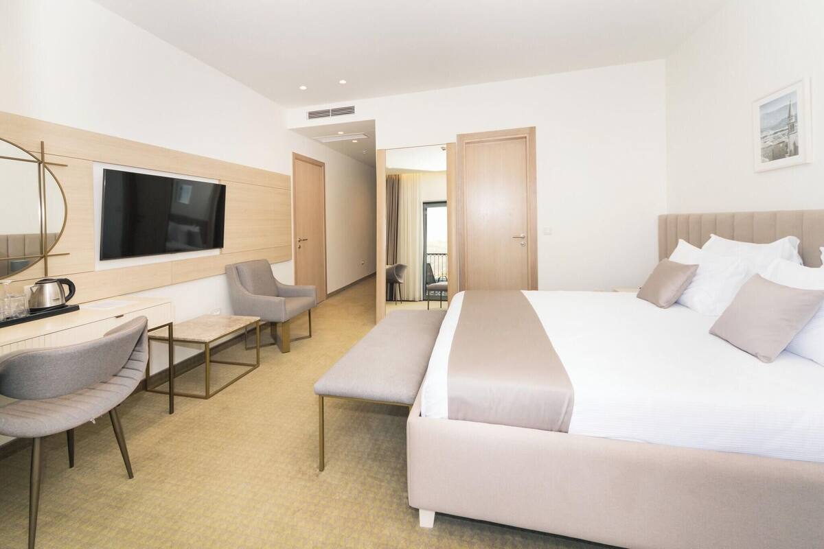 A modern hotel room with a large bed, a desk with a chair, a TV mounted on the wall, and a view of the city through the window.