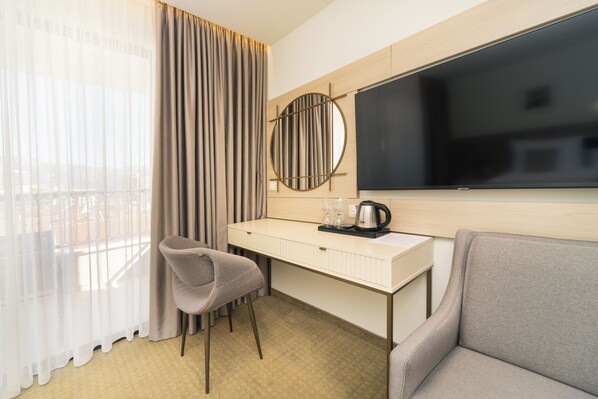 A hotel room with a desk, a chair, a sofa, a large TV, and a window with curtains. - Hotel Pima Budva (Budva)