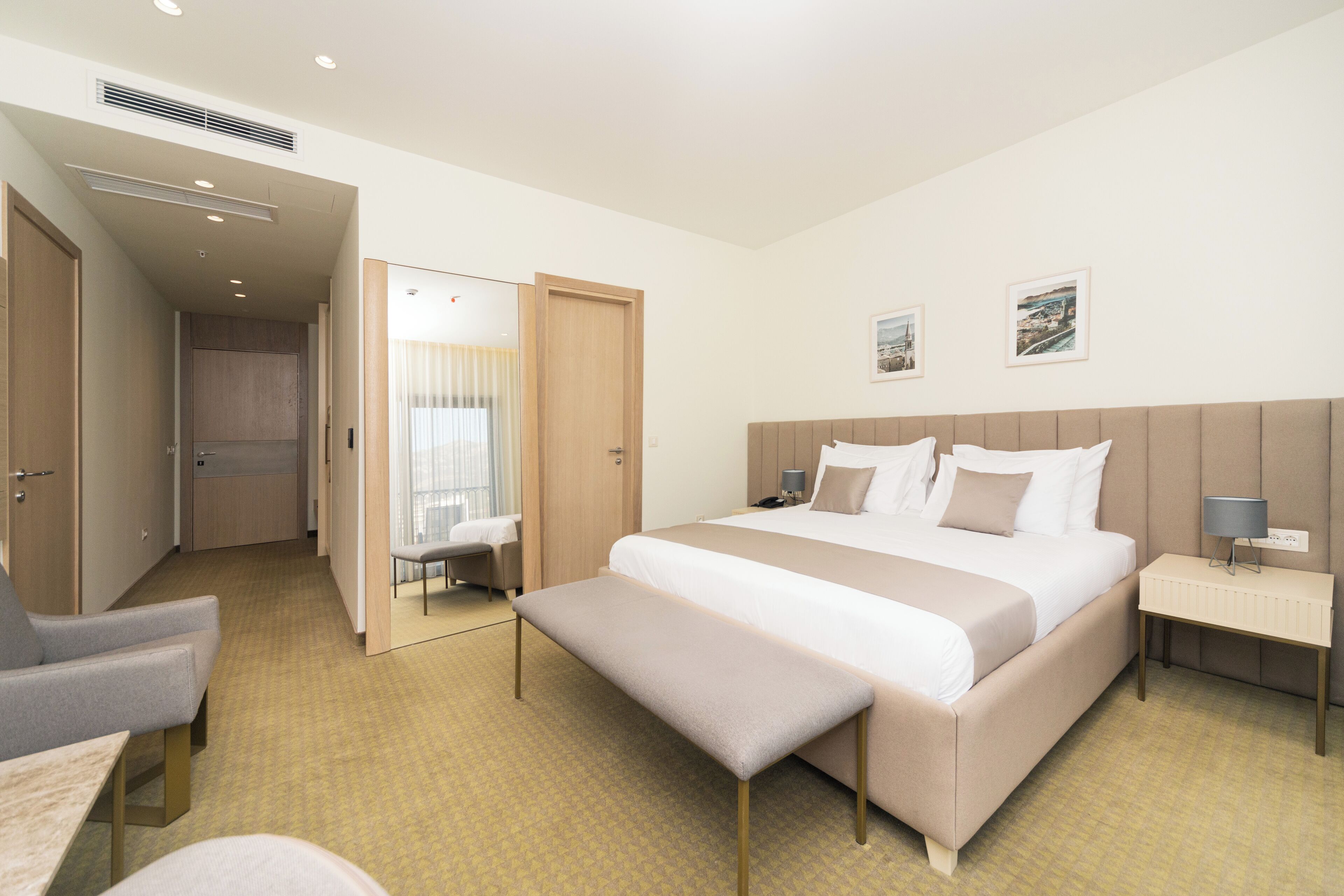 Een hotelkamer met een groot bed, een bankje, een nachtkastje met een lamp, een stoel en een bureau.