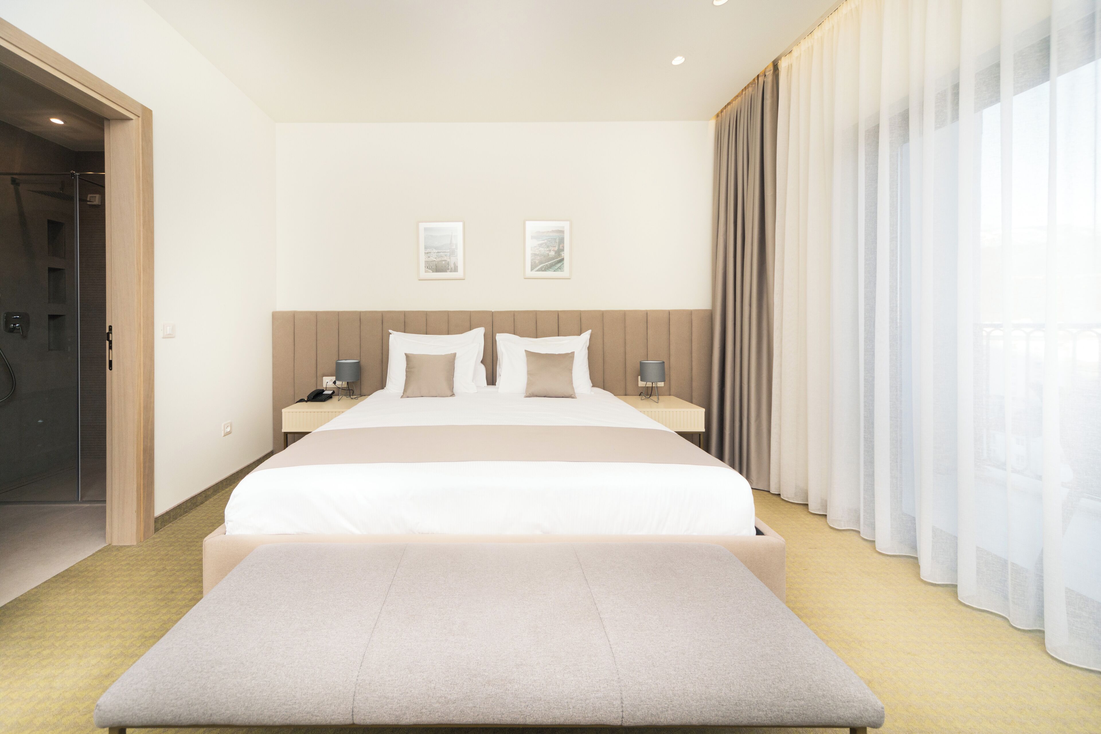 A hotel room with a large bed, bedside tables, a bench, and a view of the city through sheer curtains.