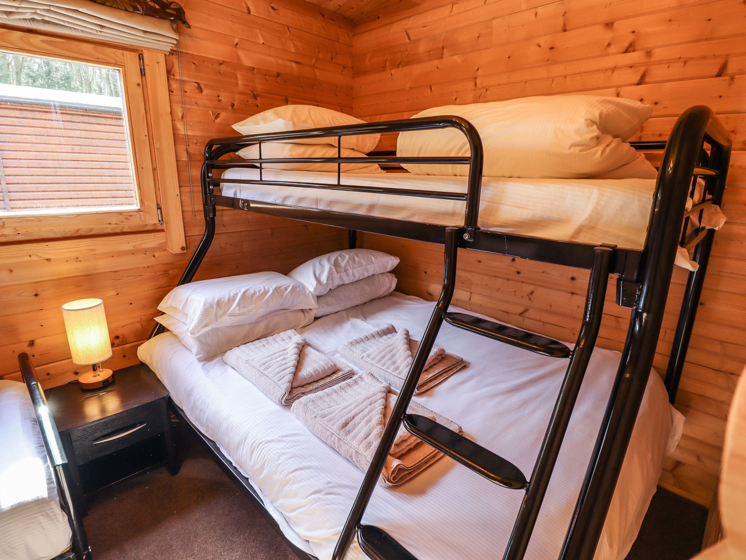 A bunk bed in a wooden cabin room with a window, a bedside table with a lamp, and a small drawer.
