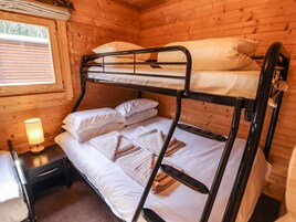 A bunk bed in a wooden cabin room with a window, a bedside table with a lamp, and a small drawer.