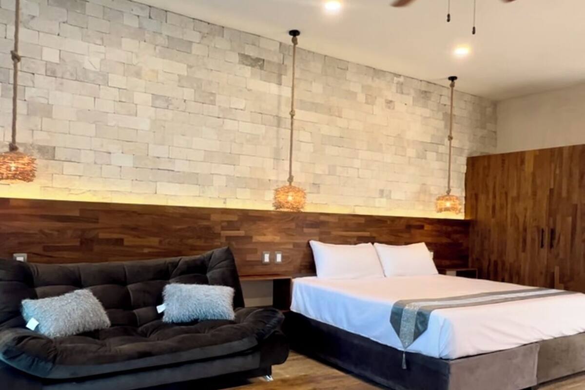 Un dormitorio moderno con pared de ladrillo, cabecera de madera, cama con ropa de cama blanca, sofá marrón oscuro con cojines y una mesa de centro de madera.