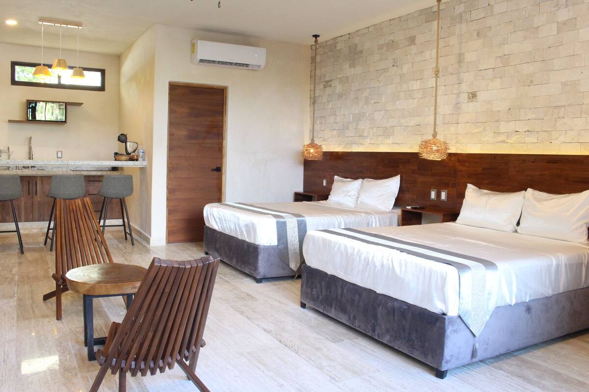 Una habitación de hotel con dos camas, una silla de madera, una mesita y una kitchenette.