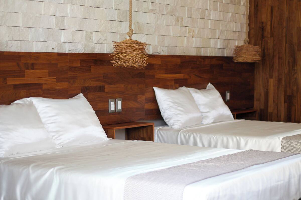 Habitación de hotel con dos camas, cabecero de madera, ropa de cama blanca y pared con textura.