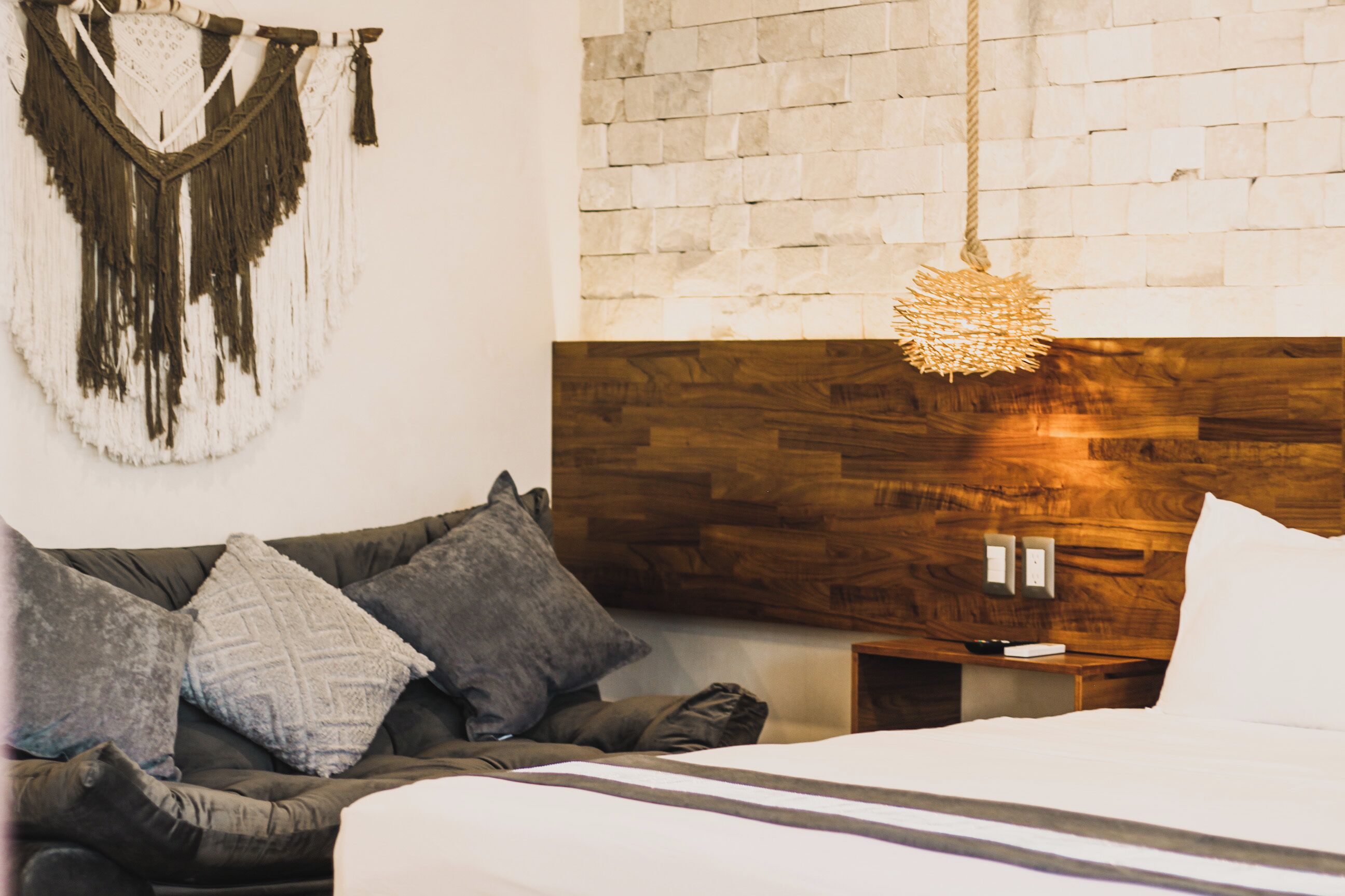 Une chambre avec une tête de lit en bois, un lit recouvert de draps blancs, un canapé gris agrémenté de plusieurs coussins et une décoration en macramé fixée au mur.