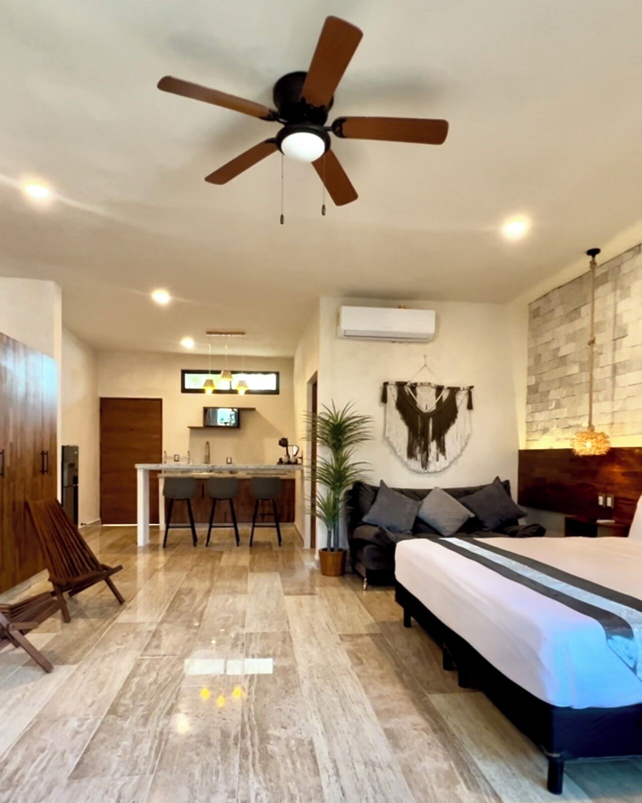 Una habitación de hotel moderna con una cama grande, ventilador de techo, comedor con cocina y sala de estar con sofá y planta.