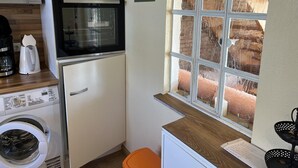 A compact kitchen with a microwave, oven, and washing machine. There's a window with a view of a brick wall. - Vacation home Deichblick (Neßmersiel)