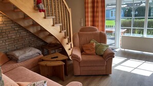 A cozy living room with a wooden staircase, a plush armchair, a sofa, and a view of the outdoors through large windows. - Vacation home Deichblick (Neßmersiel)
