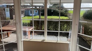 A conservatory with a view of a garden, a wooden table, and a blue house in the background. - Vacation home Deichblick (Neßmersiel)
