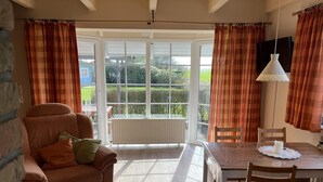 A cozy living room with a sofa, a dining table with chairs, and large windows with curtains. - Vacation home Deichblick (Neßmersiel)