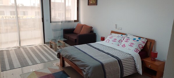 Une chambre à coucher comprenant un lit, une table de chevet, une chaise, une petite table et une fenêtre avec des rideaux légers.