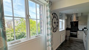 A modern kitchen with a black and white tiled floor, a large window with curtains, and a view of greenery outside. - The Market Street Apartments - Ulverston (Ulverston)