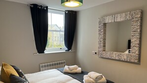 A bedroom with a bed, a window with curtains, a radiator, and a mirror with a book-themed frame. - The Market Street Apartments - Ulverston (Ulverston)