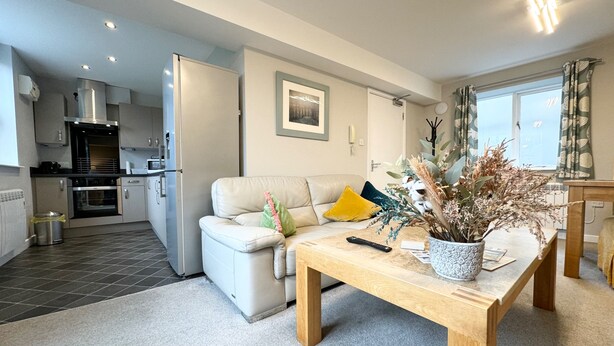 A modern kitchen with a refrigerator, oven, and microwave. There is a dining area with a wooden table and a sofa with cushions. A vase with dried flowers is on the table. - The Market Street Apartments - Ulverston (Ulverston)