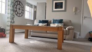 A living room with a wooden table, beige sofa, patterned curtains, a clock, and a framed picture on the wall.