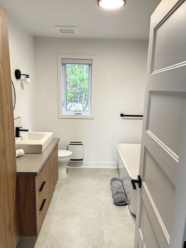 A modern bathroom with a wooden vanity, a white sink, a toilet, a bathtub, and a window with a view of trees. - The Double Diamant (St-Côme)