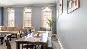 A dining area with a wooden table, chairs, and a bench. There are three arched windows with blinds, a potted plant, and framed artwork on the wall. - Lovely 2 bedroom downtown Three Rivers, sleeps 6 (Three Rivers)