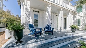 Una casa de estilo colonial blanco con un porche cubierto, dos sillas Adirondack azules y plantas en macetas.