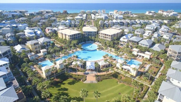Vista aérea de un complejo turístico con una piscina central rodeada de villas, un campo de golf y vistas al océano.