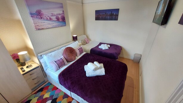 A hotel room with a bed, two bedside tables, a nightstand, a lamp, a colorful rug, and two paintings on the walls. - Ravensdale Villa- Ideal For Longer Stays in Kent (Gillingham)