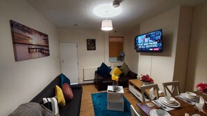 A living room with a sofa, a coffee table, a TV mounted on the wall, and a dining area with a table set for two. - Ravensdale Villa- Ideal For Longer Stays in Kent (Gillingham)