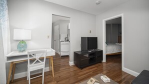 En moderne og lys stue med trægulv, et hvidt skrivebord med lampe, et sort tv-møbel, og en seng i det tilstødende rum.