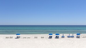 Ein Strand mit Reihen aus weißen Liegestühlen und blauen Sonnenschirmen unter blauem Himmel.