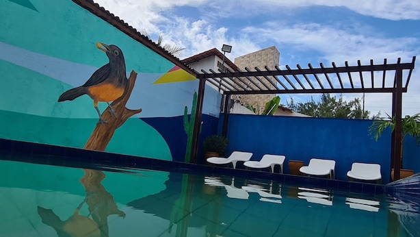 Una piscina con un mural de un pájaro en una rama, una pérgola y dos sillas reclinables blancas.