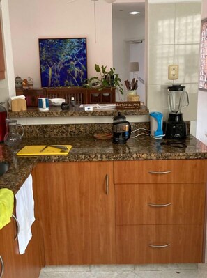 A kitchen with granite countertops, wooden cabinets, a coffee maker, a blender, and a painting on the wall.