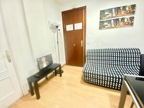 A room with a wooden door, a patterned sofa, a small table with a TV, and a floor lamp.
