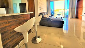 A modern kitchen with a wooden countertop and a bar stool, a living area with a sofa and a view of the outdoors, and a ceiling fan. - Near the Beach new and Modern Penthouse Coral H3 (Puerto Plata)