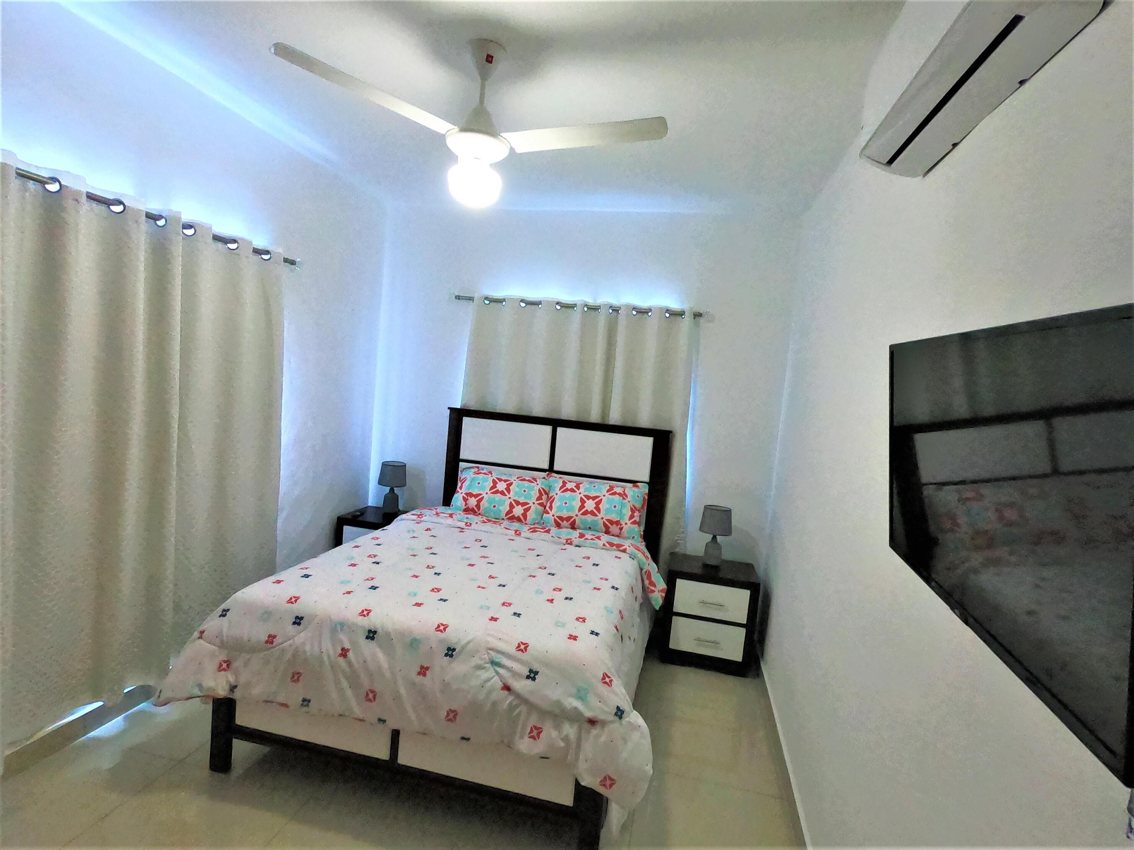 A bedroom with a bed, two bedside tables, a ceiling fan, and a window with curtains.