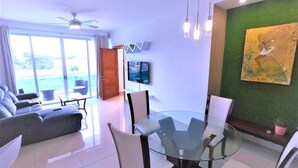 A modern living room with a glass dining table, comfortable seating, a ceiling fan, and a large window offering a view. - Near the Beach new and Modern Penthouse Coral H3 (Puerto Plata)